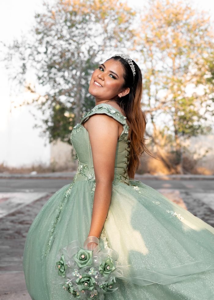 Elegant young woman celebrating quinceañera in a green gown with a tiara and bouquet outdoors.